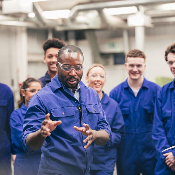 A diverse group of smiling workers in blue uniforms attentively listens to a Black man speaking and gesturing in an industrial setting.